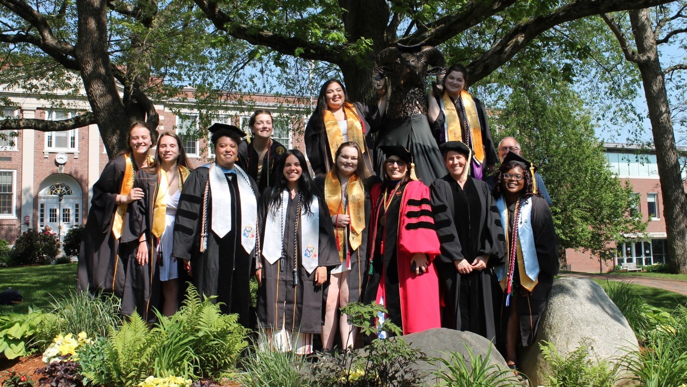 Some 2022 Biology graduates in academic regalia with faculty pose next to the Ram Statue in Crocker Grove